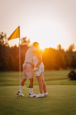 Gün batımında golf sahasında birbirine sarılan çift, yanında sarı bayrak, romantik bir atmosfer yaratıyor ve açık havada güzel bir aşk sergiliyor.