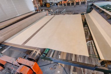 Endüstriyel bir fabrikadaki makineler üzerinde büyük rulo kağıt işleniyor. Üretim süreci ve ilgili çeşitli materyallerin dokuları sergileniyor.