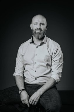 Sakallı, açık renk gömlekli, koyu renk kotlu koyu renk pantolonlu, koyu renk bir arka plana karşı tabureye oturmuş, çarpıcı bir monokrom portresi yaratıyor.