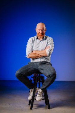 Sakallı, kendine güvenen kel bir adam modern bir stüdyoda, mavi arka planda bir taburede oturuyor. Rahatlamış atmosferi arttırıyor ve kendine güvenen varlığını vurguluyor.