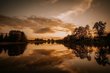 Bereketli yeşilliklerle çevrili durgun suya yansıyan canlı renkler içeren sakin günbatımı manzarası huzurlu ve huzurlu bir ortam yaratıyor.