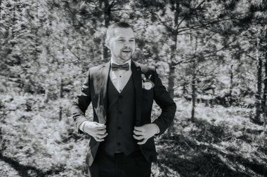 Siyah takım elbiseli şık bir adam yeşil ile çevrili canlı bir ormanda kıyafetlerini ayarlar. Doğada bir karmaşa ve cazibe anı yakalar.