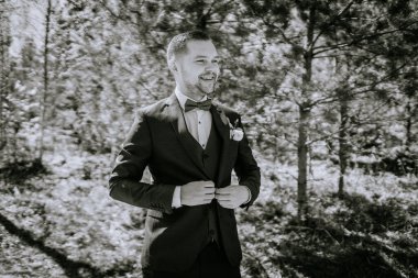 Siyah takım elbiseli adam dışarıda papyonunu ayarlıyor. Ağaçlar ve yeşilliklerle çevrili. Düğün atmosferinde mutluluk ve incelik yayıyor.