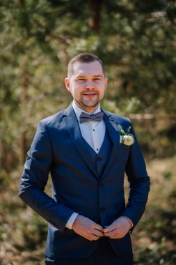 Resmi takım elbiseli kendine güvenen bir adam açık havada poz verir, yemyeşil, sakin bir ortamda zarafet ve stili ön plana çıkarır.