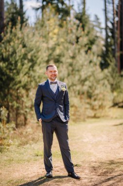 Resmi giyinmiş bir erkek, ağaçlar ve yeşilliklerle çevrili sakin bir açık havada duruyor. Doğal bir ortamda kendine güven ve tarz yayıyor.