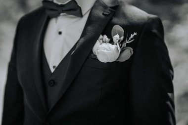 Siyah takım elbiseli, papyonlu şık bir adam, beyaz bir çiçek yaka çiçeğiyle süslenmiş, resmi bir atmosferde zarafet ve cazibe yayıyor.