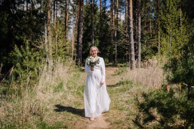 Zarif beyaz elbiseler içindeki gelin, manzaralı bir orman yolunda yürüyor, güzel bir buket tutuyor, canlı yeşil ve uzun ağaçlarla romantik bir atmosfer yaratıyor.