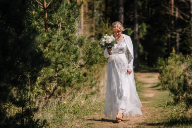 Zarif beyaz elbiseli gelin orman yolunda yürüyor, elinde güzel bir buket tutuyor, canlı yeşillikler ve uzun ağaçlarla çevrili, huzur ve romantizmi çağrıştırıyor.