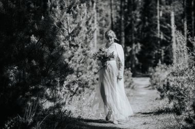 Beyaz elbiseli bir gelin, ağaçlar ve yeşilliklerle çevrili bir çiçek buketini tutarak orman yolunda yürüyor. Huzurlu bir düğün atmosferini çağrıştırıyor.