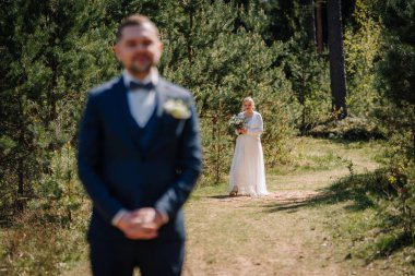 Resmi damat manzaralı bir orman yolunda dururken zarif beyaz elbiseli gelin canlı yeşilliklerle çevrili buketi tutuyor ve romantik bir atmosfer yaratıyor.
