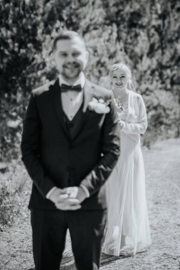 Resmi damat ellerini sıkarak ayakta duruyor, zarif beyaz elbiseli gelin arkadan yaklaşıyor, aşkla dolu güzel bir açık hava düğün sahnesi yaratıyor.