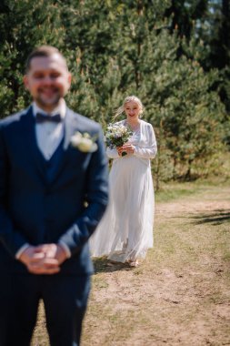 Beyaz elbiseli neşeli gelin, damada resmi giysiler içinde yaklaşır, canlı yeşillik zeminine karşı kurulur, romantik bir açık hava düğün atmosferi yaratır.