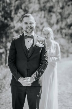 Resmi damat siyah takım elbise içinde gülümserken, gelin beyaz elbiseyle yaklaşıyor, doğal bir zemin üzerine kuruluyor, neşeli bir düğün ortamı yaratıyor.