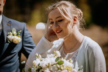 Açık renk elbisesi ve bir buket beyaz çiçekle gülümseyen gelin, yemyeşil çiçeklerle çevrili, güzel bir düğün töreninde neşe ve aşkı simgeliyor.