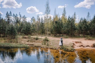 Romantik çift sakin bir su gövdesiyle samimi bir anı paylaşıyor, arka planda canlı yeşillikler ve ağaçlar, barışçıl bir açık hava ortamı yaratıyor.