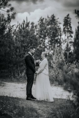 Gelin ve damat, sakin bir ormanda, ağaçlarla çevrili bir buketle, doğal bir çevrede sevgi ve birliği simgeleyen zarif kıyafetler içinde dururlar.