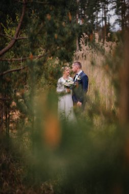 Gelin ve damat, yemyeşil ve uzun otlarla çevrili sakin bir açık hava sahnesinde kucaklaşıp özel günleri için romantik bir ortam yaratıyorlar.