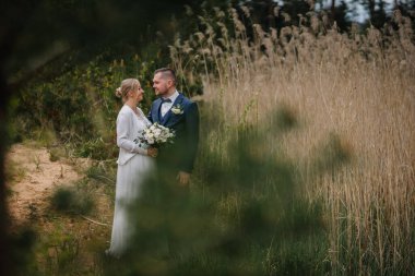 Gelin ve damat zarif bir gelinlik içinde, doğal bir manzarada yan yana duruyorlar, uzun otlarla çevrili, güzel bir ortamda aşklarını ve neşelerini sergiliyorlar.