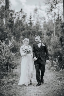 Resmi nikah kılığındaki bir çift, yemyeşil ormanlarla çevrili bir yolda birlikte yürürken doğada bir neşe ve yakınlık anı yakalıyorlar.