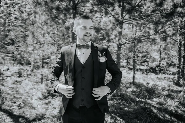 Siyah takım elbiseli şık bir adam yeşil ile çevrili canlı bir ormanda kıyafetlerini ayarlar. Doğada bir karmaşa ve cazibe anı yakalar.