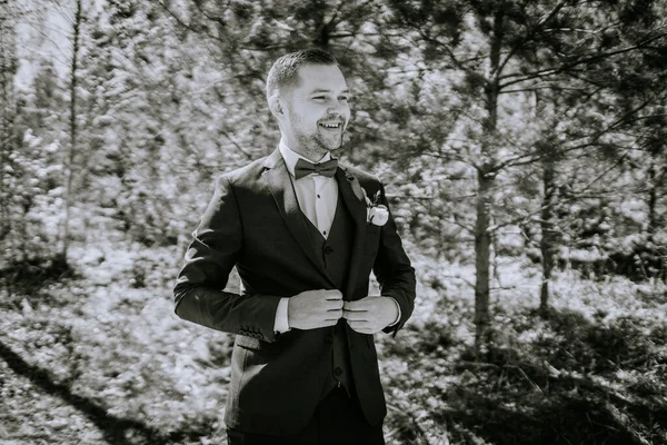 Siyah takım elbiseli adam dışarıda papyonunu ayarlıyor. Ağaçlar ve yeşilliklerle çevrili. Düğün atmosferinde mutluluk ve incelik yayıyor.