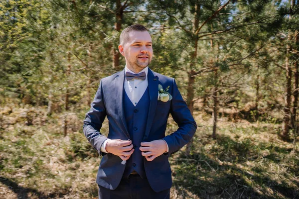 Donanma kıyafeti giymiş bir adam ağaçların arasında duruyor. Kendine güveni ve tarzı var. Düğün teması ya da resmi etkinlikler için ideal.
