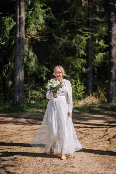 Zarif beyaz gelinlik içinde, neşeyle gür ormanda yürüyor, elinde çiçekler buketi, yeşillik ve güneş ışığıyla, romantizm ve doğa duygusunu çağrıştırıyor.