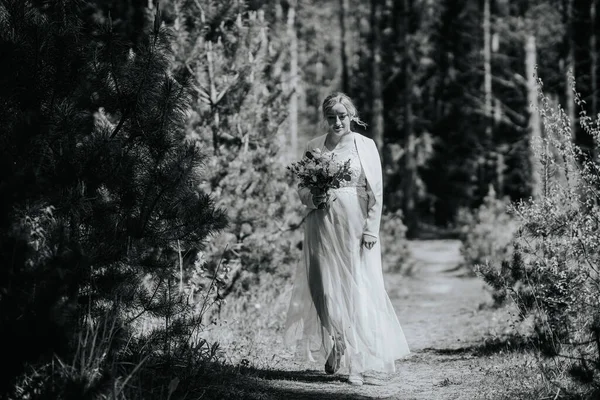 Beyaz elbiseli bir gelin, ağaçlar ve yeşilliklerle çevrili bir çiçek buketini tutarak orman yolunda yürüyor. Huzurlu bir düğün atmosferini çağrıştırıyor.