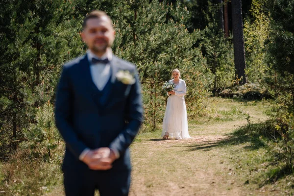 Resmi damat manzaralı bir orman yolunda dururken zarif beyaz elbiseli gelin canlı yeşilliklerle çevrili buketi tutuyor ve romantik bir atmosfer yaratıyor.