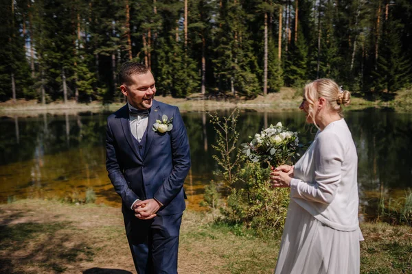 Damat resmi giysiler içinde, doğadaki sevgi ve kutlamayı yansıtan canlı yeşilliklerle çevrili, huzurlu bir gölün kenarında annesiyle neşeli bir anı paylaşıyor.