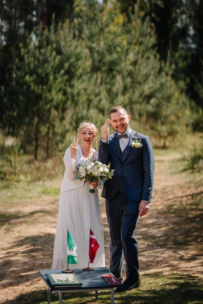 Mutlu yeni evliler düğünlerini açık hava ortamında kutluyorlar, el ele tutuşuyorlar ve barış işaretleri yapıyorlar, masanın üzerinde bir buket ve bayraklar, neşe ve sevgi yayıyorlar.