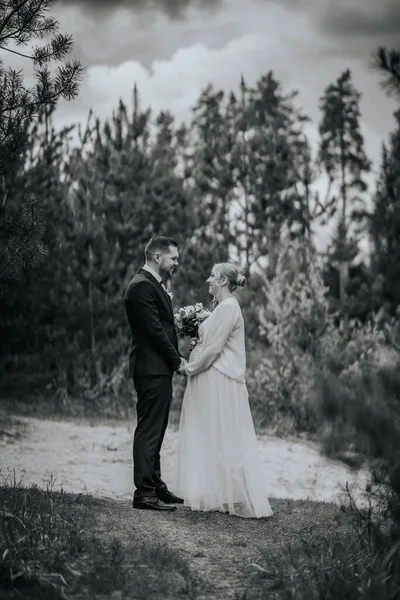 Gelin ve damat, sakin bir ormanda, ağaçlarla çevrili bir buketle, doğal bir çevrede sevgi ve birliği simgeleyen zarif kıyafetler içinde dururlar.