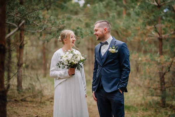 Düğün kıyafetli bir çift, sakin bir ormanda duruyor, gülüşüyorlar, yeşillikler ve ağaçlar özel anları için romantik bir ortam yaratıyorlar.