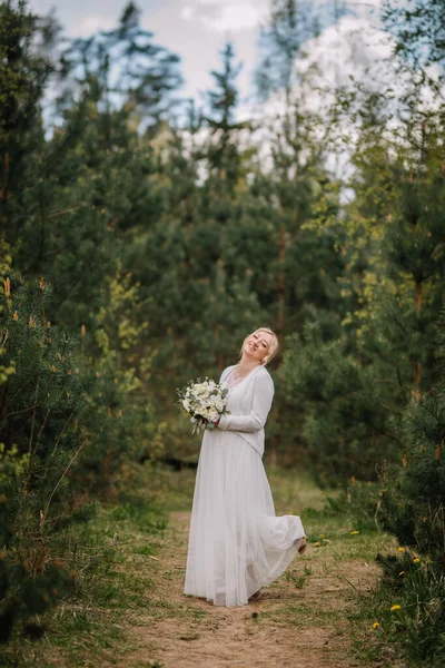 Zarif beyaz elbiseler içindeki neşeli gelin, elinde buket çiçekle, yeşil ve yumuşak ışıkla romantik ve büyüleyici bir atmosfer yaratıyor.