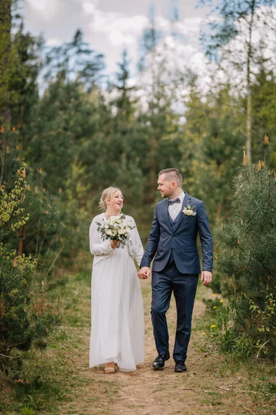 Düğün kıyafetli bir çift birlikte orman yolunda yürüyor, yeşilliklerle çevrili, doğanın güzelliğinde aşkın ve kutlamanın özünü yakalıyorlar.