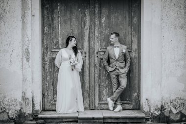 Düğün kıyafetleri içinde çift, romantik ve zarif bir şekilde, kırsal ahşap kapıların yanında durur. Yumuşak ışıklandırma, özel günlerinin neşeli atmosferini güçlendirir.