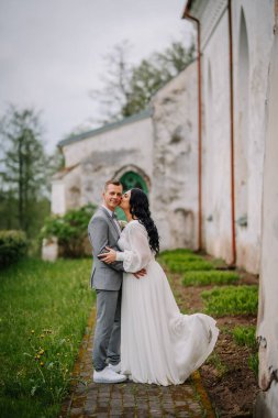 Düğün kıyafetli çift, arka planda yeşillik ve eski mimariyle romantik ve neşeli bir atmosfer yaratan açık havada sevgi dolu bir anı paylaşıyor.