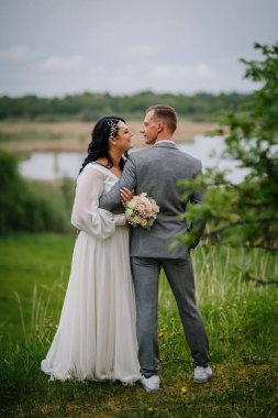 Düğün kıyafetli çift doğada hassas bir anın tadını çıkarıyor. Arka planda yeşillik ve su, güzel bir ortamda sevgi ve birliği simgeliyor.