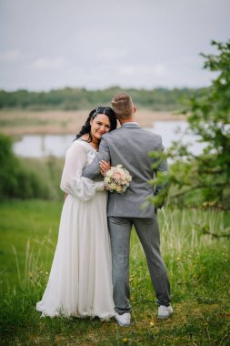 Beyaz elbiseli zarif gelin, gri takım elbiseli damadı kucaklıyor, yeşil ve sakin suların arka planına kuruluyor, dışarıda güzel bir düğün atmosferi sergiliyor.