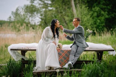 Çift, yeşil bir manzarada piknik masasında neşeli bir anın tadını çıkarıyor. Zarif bir şekilde giyinmiş, doğada aşkın ve kutlamanın özünü yakalıyor.