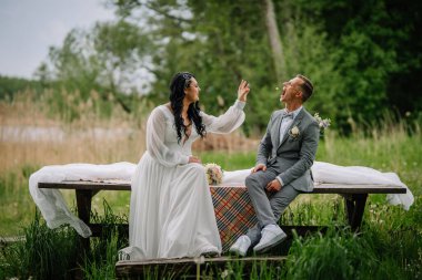 Düğün için giyinmiş çiftler, doğadaki kırsal bir bankta eğlenceli etkileşimlerden zevk alıyorlar, yeşil ve yumuşak ışıklarla çevrili, romantik bir ortam yaratıyorlar.