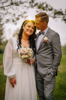 Düğün kıyafeti giymiş, gülümseyen ve doğa ile çevrili sakin bir ortamda kucaklaşan, resim gibi bir ortamda sevgi ve kutlama sergileyen bir çift.