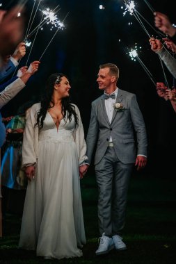 Mutlu çift el ele yürür, misafirlerin tuttuğu havai fişeklerle aydınlatılır, güzel bir düğün gecesi kutlamasında romantik bir ortam yaratır.