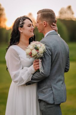 Düğün kıyafetli çift, ellerinde güzel bir buketle dışarıda hassas bir anı paylaşıyorlar, yemyeşil ve sıcak güneş ışığı altında romantik bir atmosfer yaratıyorlar.