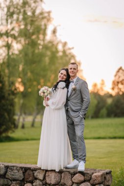 Düğün kıyafetleri giymiş çift taş duvarda poz veriyor. Canlı bir günbatımı ve arka planda yeşillik var. Özel günleri için romantik bir ortam yaratıyorlar.