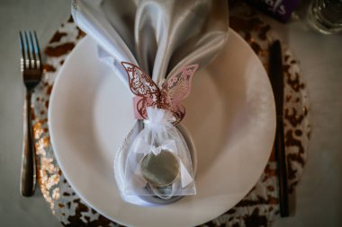 Masa sehpaları, bir kelebek, dekoratif bir hediye çantası ve cilalanmış çatal bıçaklarla süslenmiş zarif bir peçete, sofistike bir yemek atmosferi yaratıyor.