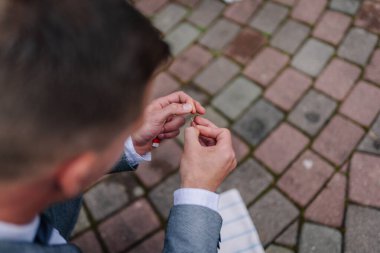 Gri takım elbiseli bir erkek sigara yuvarlıyor. Kaya kaldırımlarında. Açık havada bir an için odak ve beceri gösteriyor.