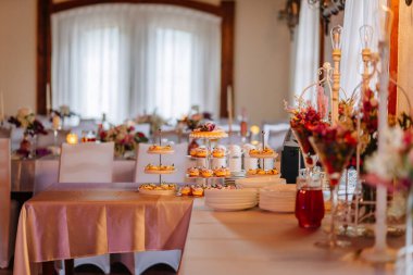 Çiçekli süslemeler ve hafif ışıklandırmalarla çevrili bir tatlı masasını çok güzel düzenlenmiş bir ziyafet salonu, neşeli bir ortam yaratıyor.