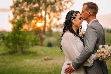 Çift gün batımında hassas bir anı paylaşıyor, arka planda yemyeşil bir ortam, gelin buketi tutuyor, kutlama için romantik ve neşeli bir ortam yaratıyor.