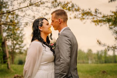 Düğün kıyafetli çift açık havada samimi bir anı paylaşıyor, yemyeşil ve yumuşak doğal ışıkla çerçevelenmiş, sevgi ve neşe duyguları uyandırıyor.